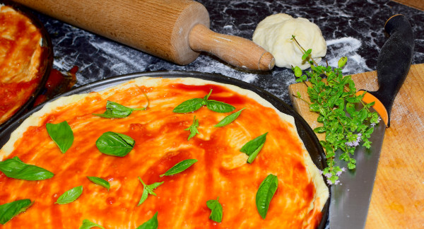 A pizza in preparation with marinara sauce and basil leaves. A rolling pin and a knife are in the background